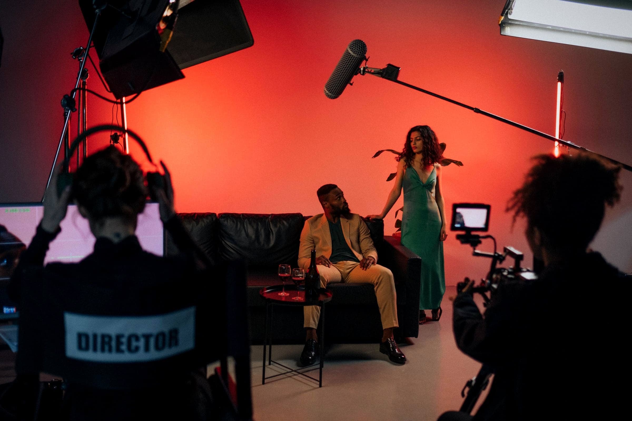 A camera operator and crew shoot a scene on a film set lit in deep red, actors on a couch in frame.